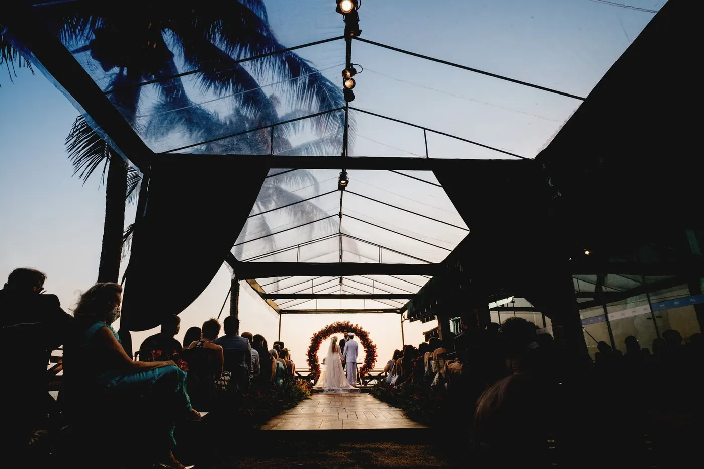 An evening wedding ceremony under a glass tent lit with warm lights and greenery