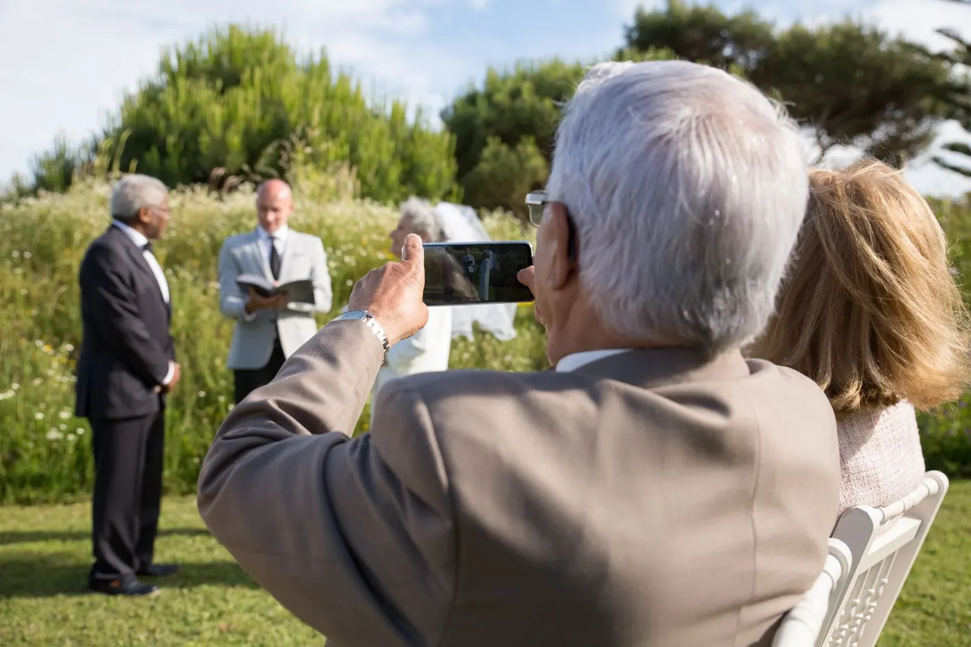 An elderly guest raising a phone to photograph an outdoor wedding ceremony