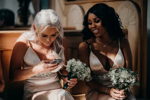 Two bridesmaids holding bouquets, smiling at a phone.