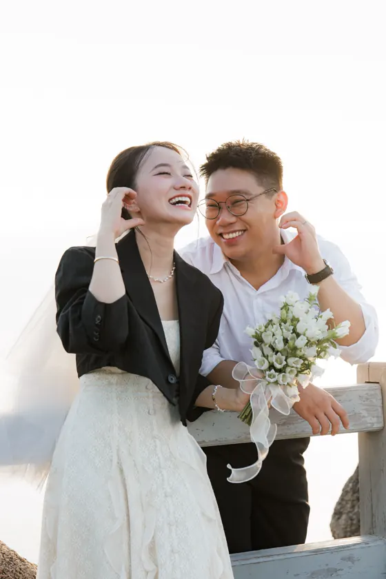 A laughing couple outdoors, the bride holding white flowers.